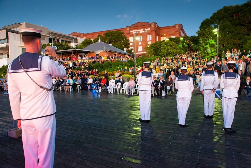 A bugler plays in front of four navy officers on parade for a dawn service in front of a large crowd in Teneriffe in Brisbane