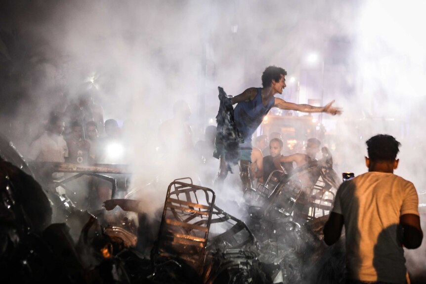A man stands on top of burnt car wreckage with an outstretched hand outside a Cairo cancer hospital