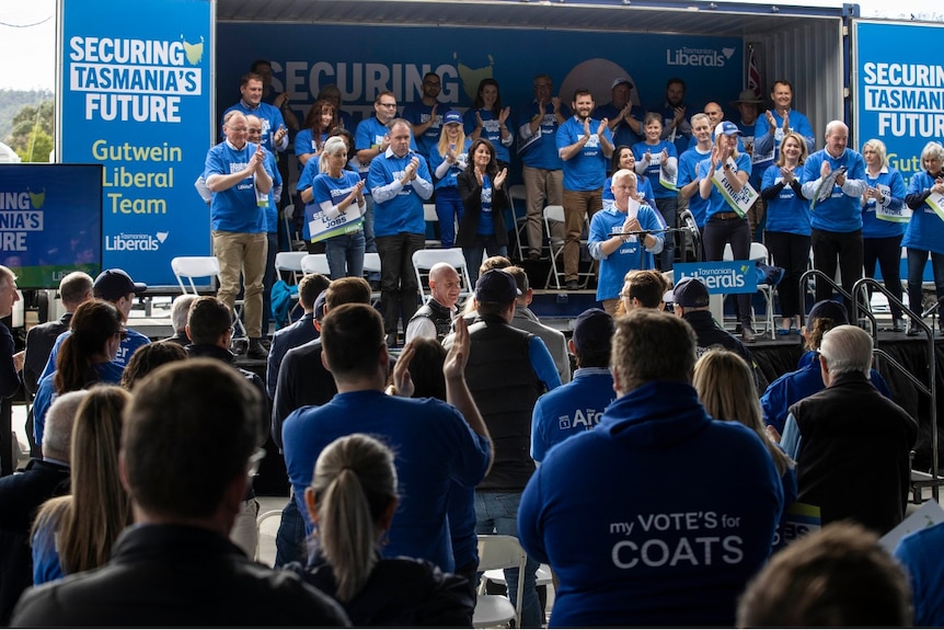 A group of people in blue t-shirts applaud a bald man in a crowd at an election rally