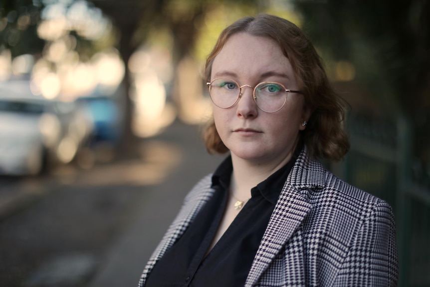 A woman wearing glasses stands on a footpath, looking at the camera with a neutral expression on her face.