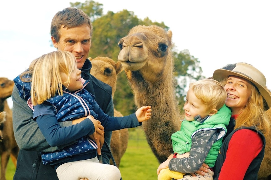 A man and woman each holds a child as they smile looking at the camera, a camel stands between them.  