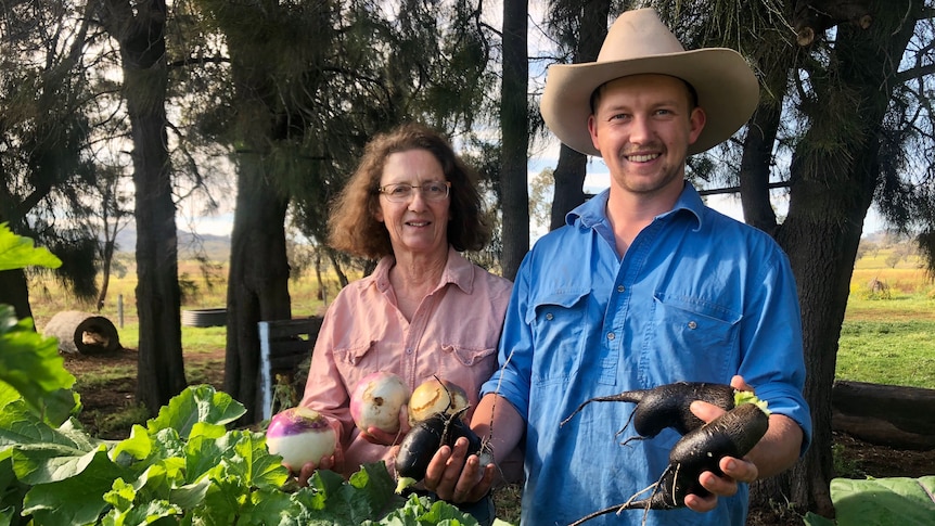 Woman in pink collared shirt and man in blue collared shirt smile and hold turnips, with trees in background.