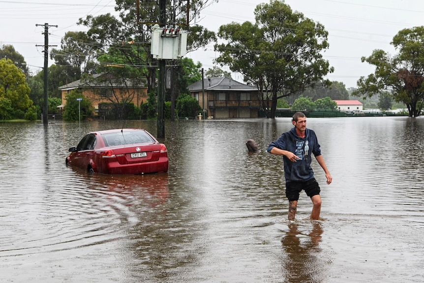 A man walks through floodwater, a partially submerged car in the background.