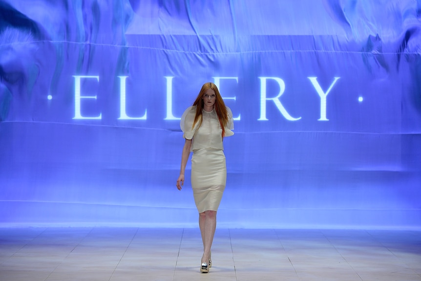 A model wearing white walks along a catwalk behind a purple background that reads ELLERY