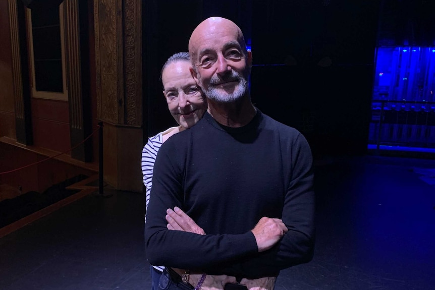 A woman puts her arms around her husband's waist as they pose for a photo on a theatre stage