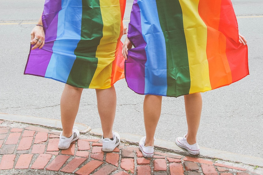 Tow people wearing white sneakers and wrapped in rainbow pride flags stand on a bricked sidewalk