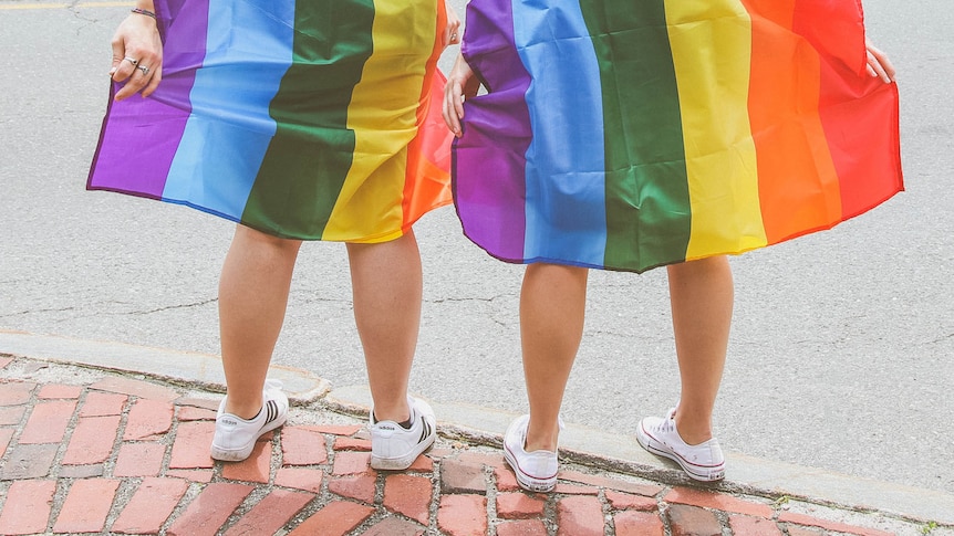 Tow people wearing white sneakers and wrapped in rainbow pride flags stand on a bricked sidewalk