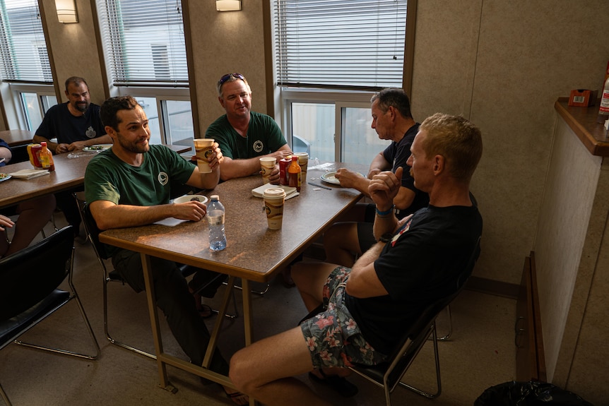 Four men sit around a table in a dining hall with cups of tea, laughing, the afternoon light hitting their faces.