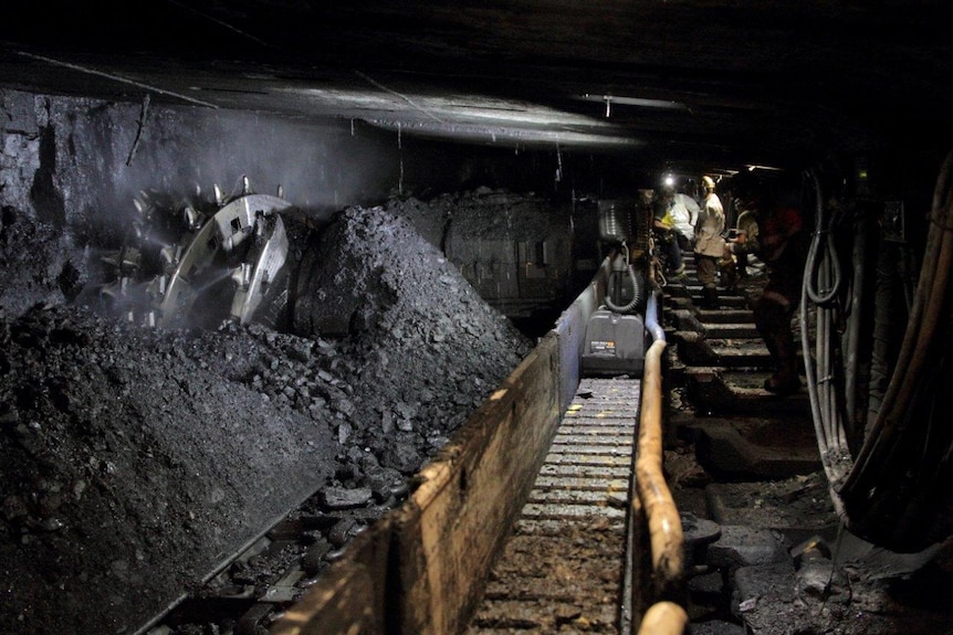 The interior of a longwall mineshaft. A drill, conveyor belt and miners wearing headlamps are visible.
