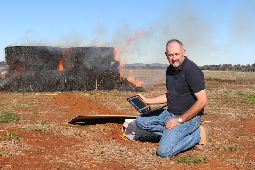 A man in a blue shirt kneels in front of a burning haystack