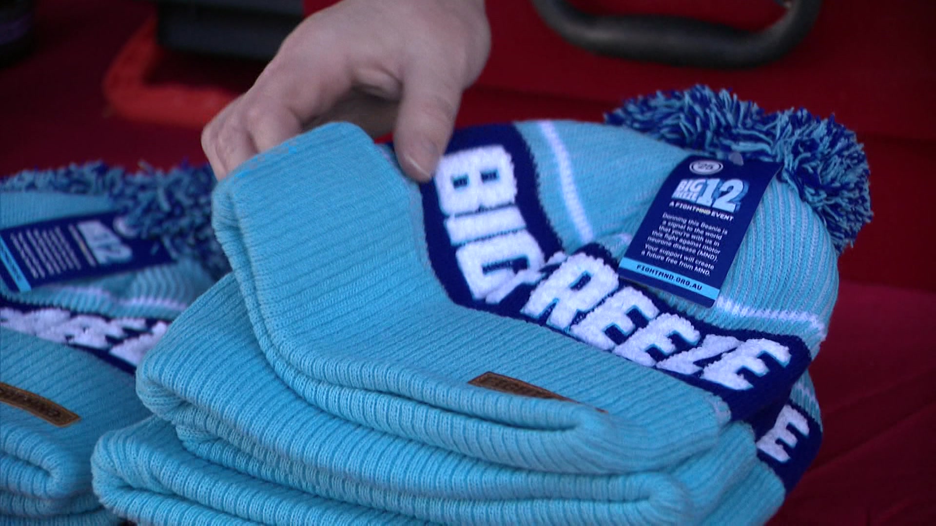 A hand picks up a light and dark blue beanie that says "Big Freeze" from a stack of other beanies.