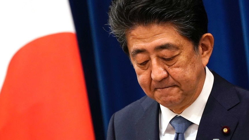 Shinzo Abe looks down as he speaks in front of a Japanese flag.