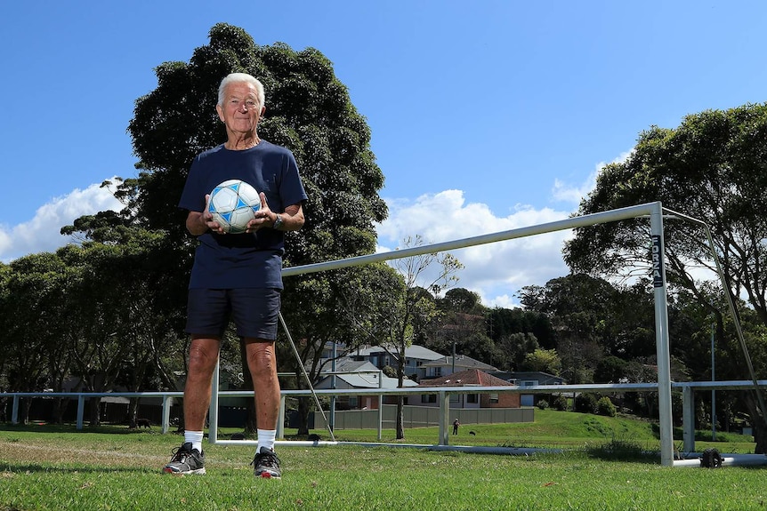 Peter Webster holds a blue and white soccer ball in front of goal posts on a soccer field.