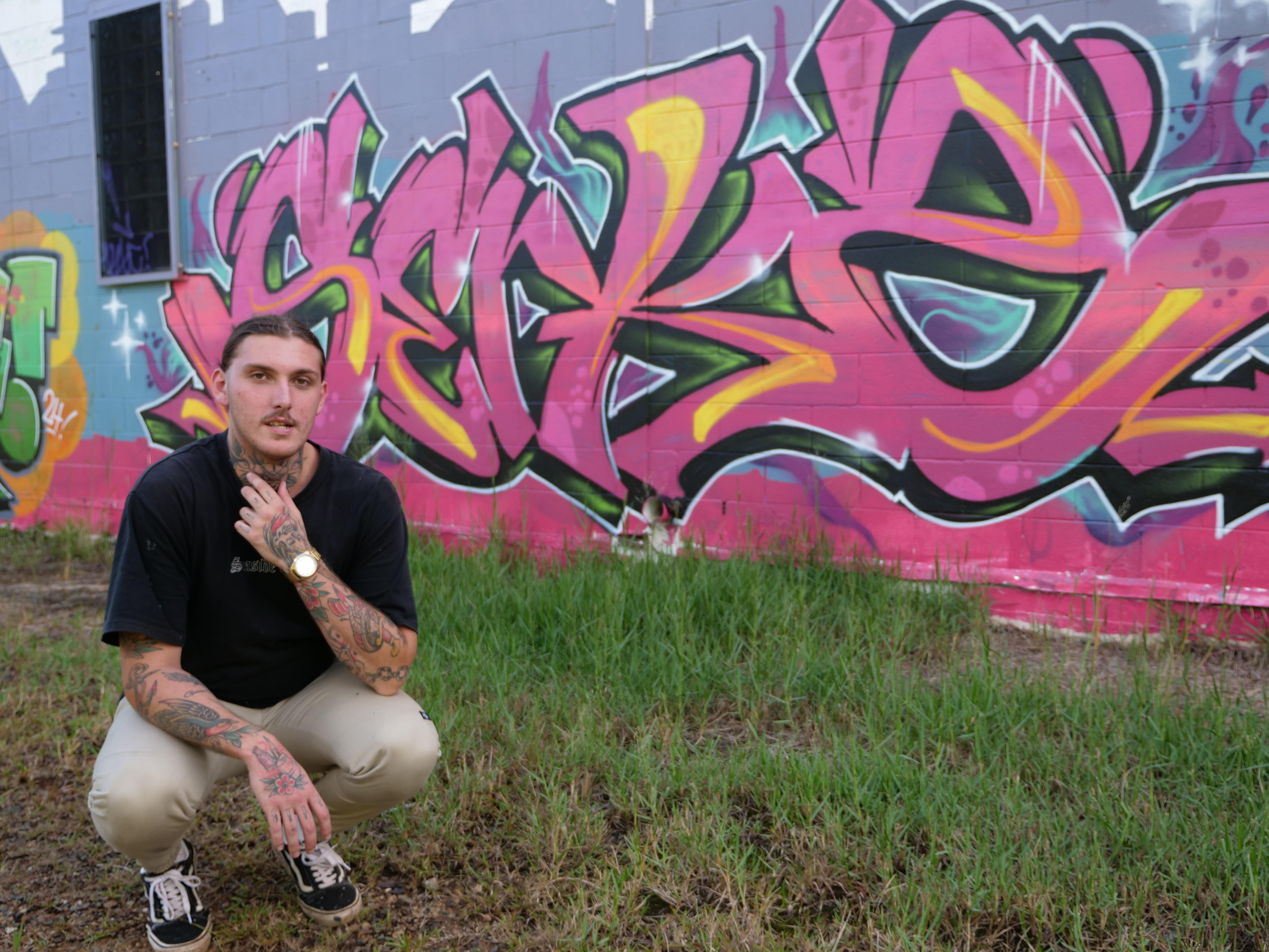 A young man with tattoos and long hair pulled back, crouched on grass before a large pink and yellow graffiti word on a wall 