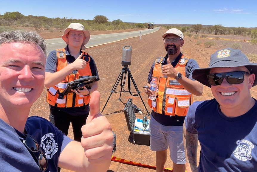 Four men stand on the side of a remote road with their thumbs up and radiation equipment in the background