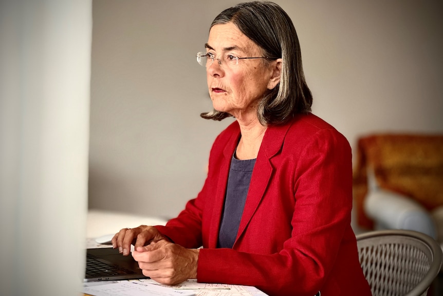 Jenny Buchan sits at a desk wearing a red jacket, she is a middle-aged woman with rimless glasses and shoulder-length hair.