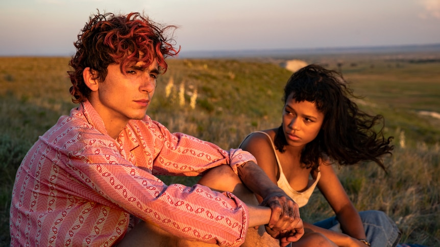 A pale young man with dyed red hair and a young Black woman sit together at sunset, him looking into distance