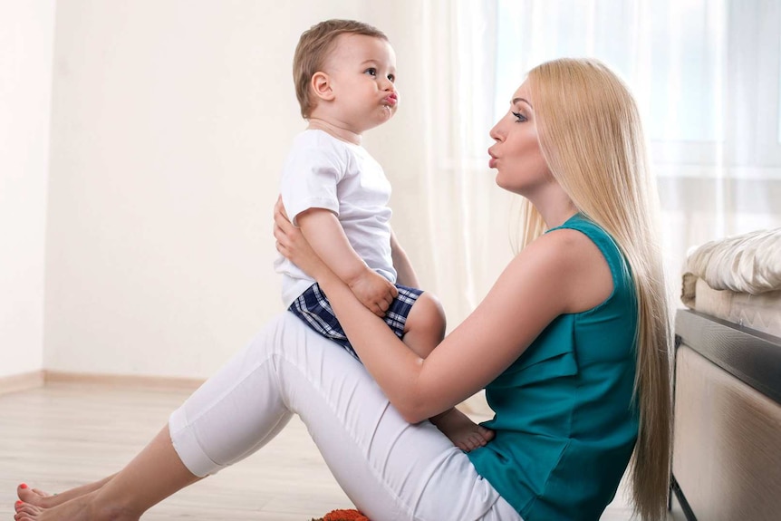 A blonde mother sits on the floor with her baby on her knees.