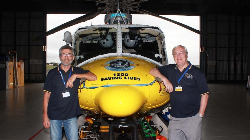 Two men on either side of a yellow and blue helicopter smiling