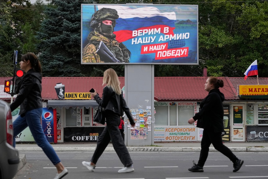 People walk past a billboard displaying a soldier and a Russian flag.