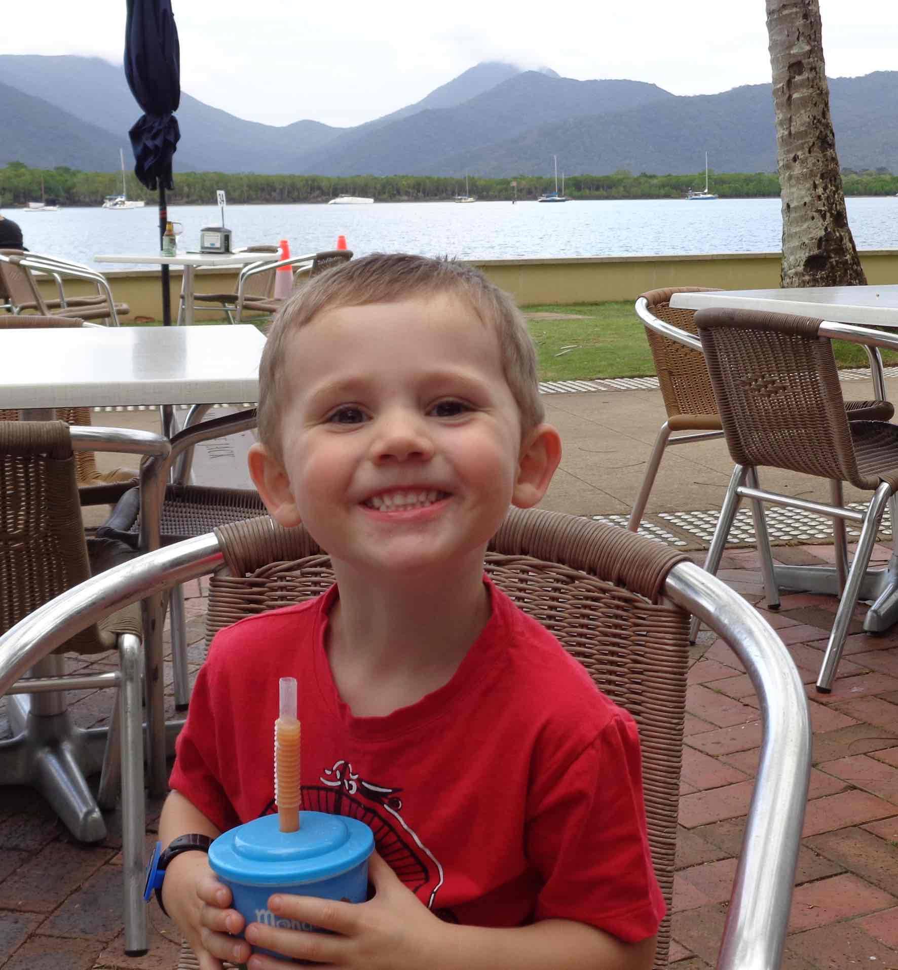 a young boy smiling at the camera while sitting down outdoors and holding a sippy cup