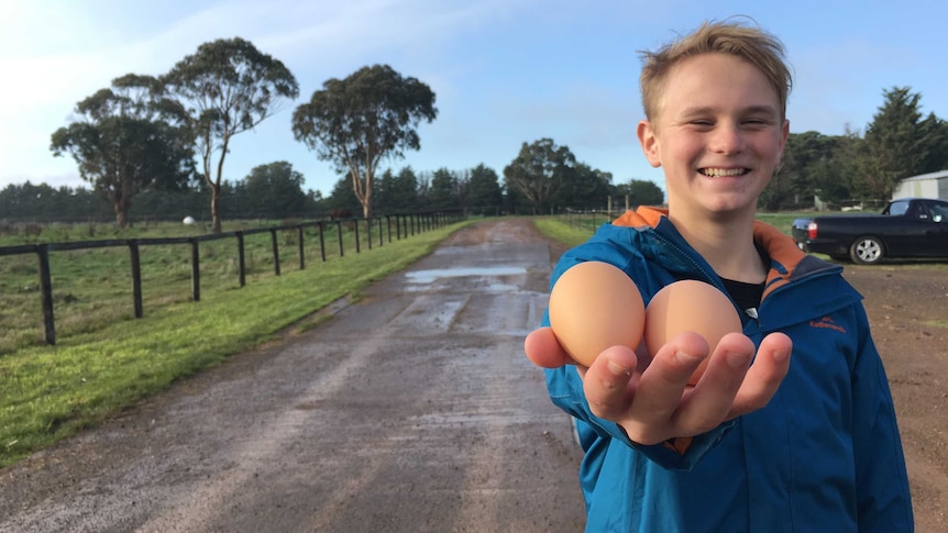 A young man who is smiling stands with his hand lifted toward the camera holding two brown eggs