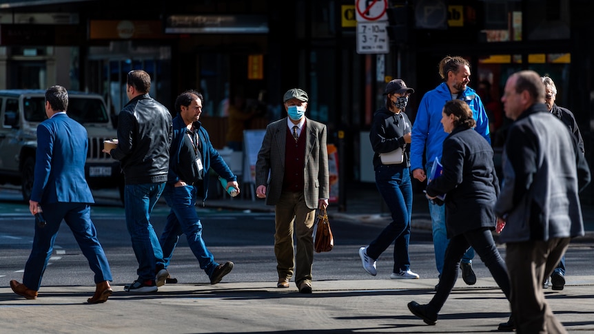 Pedestrians crossing a city road in all different directions. A man walking toward the camera wears a blue surgical mask