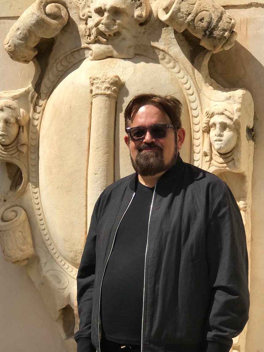 Man with dark hair, beard and glasses stands against a cultural background