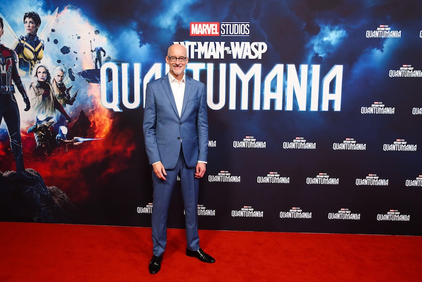 Man in blue suit smiles while standing in front of movie poster for Ant-Man