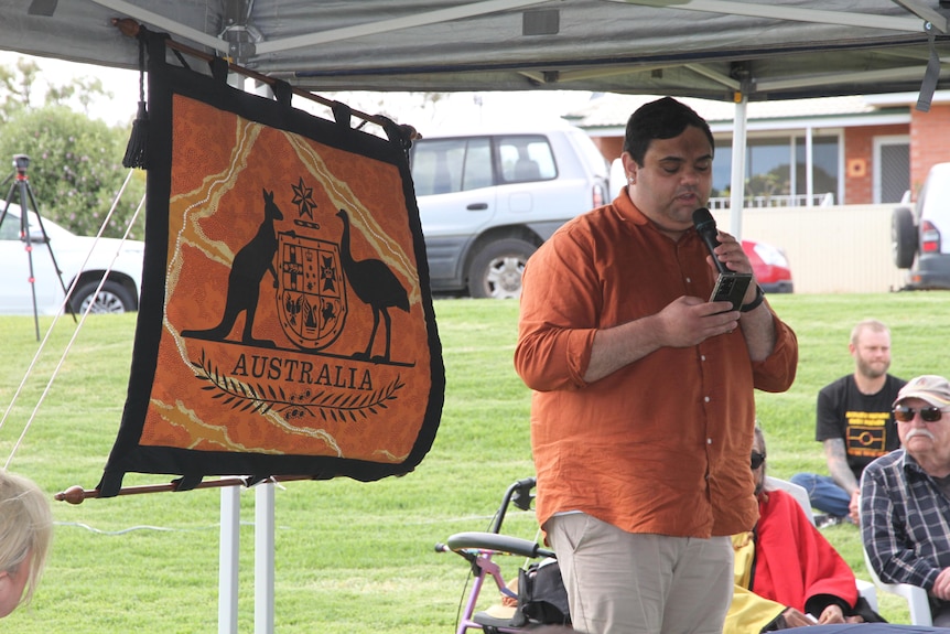 A young Indigenous man wears orange next to an orange sign that says Australia speaking on the microphone. 