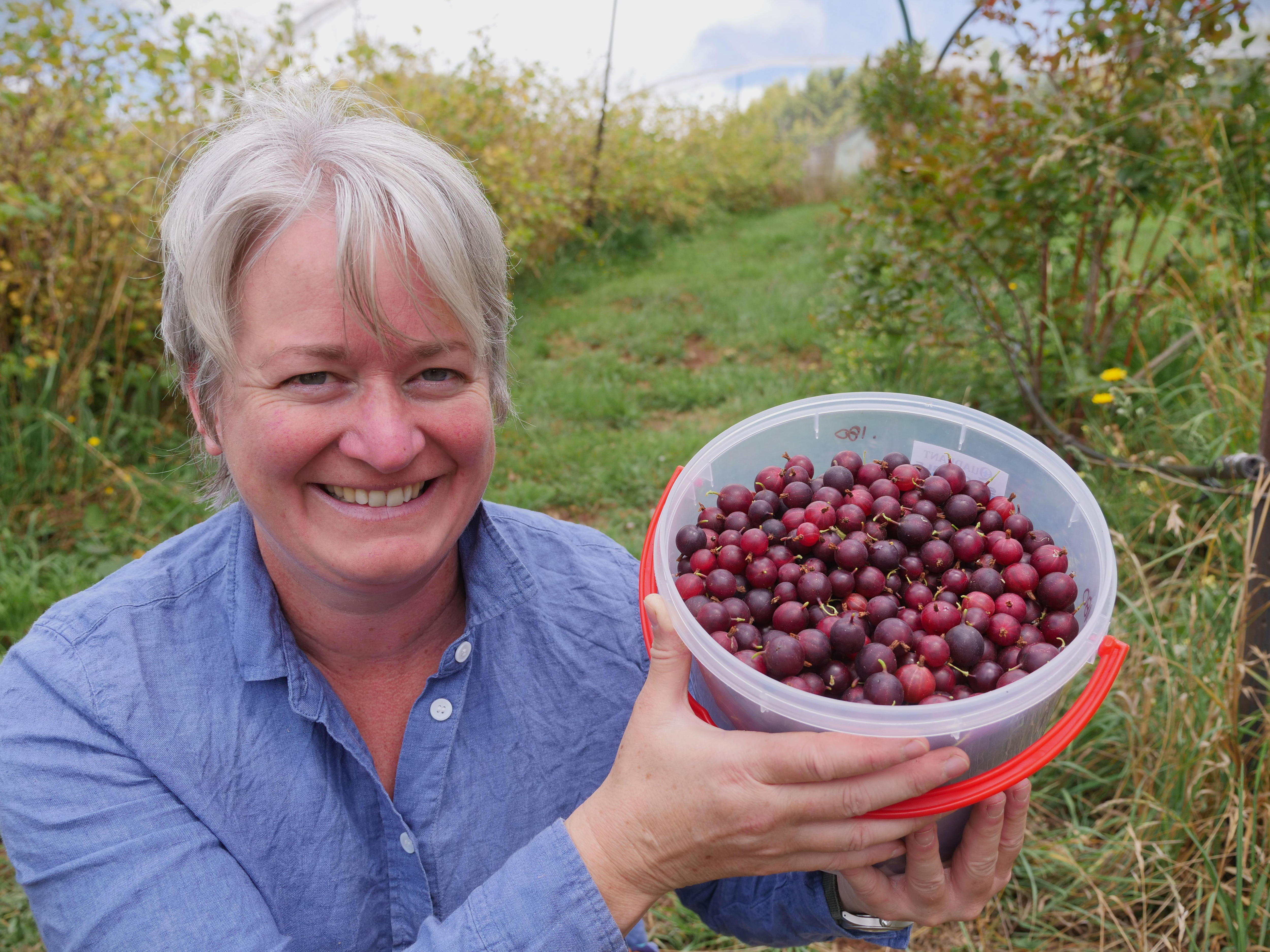 A woman with short hair smiling as she holds up a bucket filled with gooseberries.
