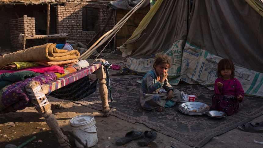 Two young Afghan girls eat on the ground in a run-down block of land.