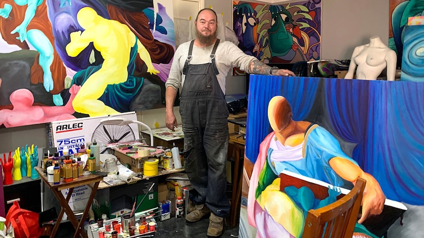 Artist Russell Sanderson stands in the centre of a room surrounded by large, brightly coloured canvases.