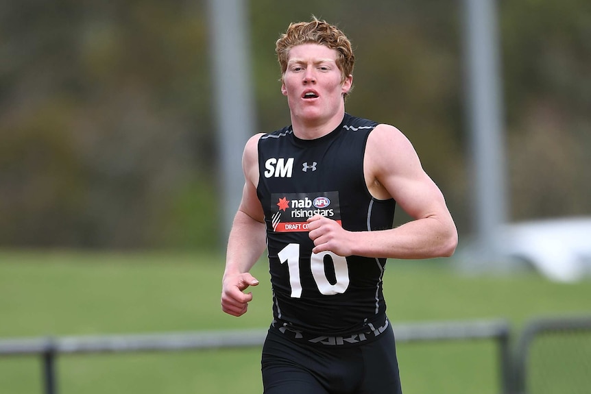 A male Australian rules footballer wearing a singlet runs around a track.