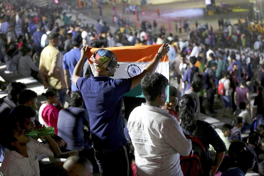 A crowd of people. One of them is a man holding up an Indian flag