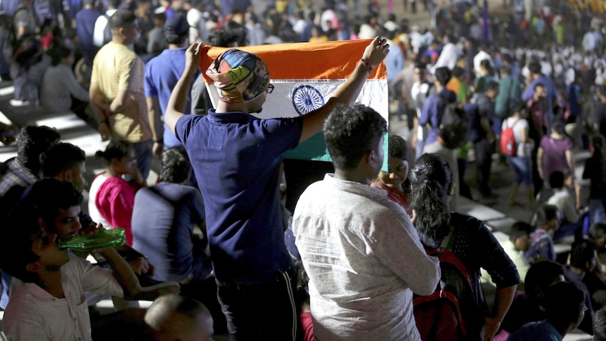 A crowd of people. One of them is a man holding up an Indian flag