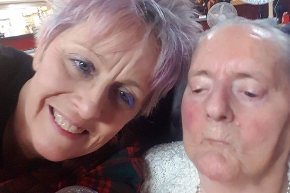 An older woman with purple hair poses for a photo with her elderly mother.