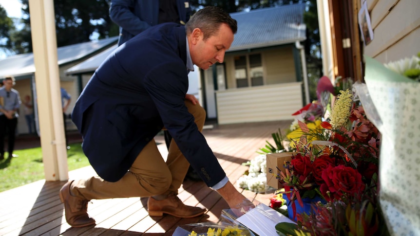 The premier crouches forward to place flowers along a wall where there are more flowers on a verandah.