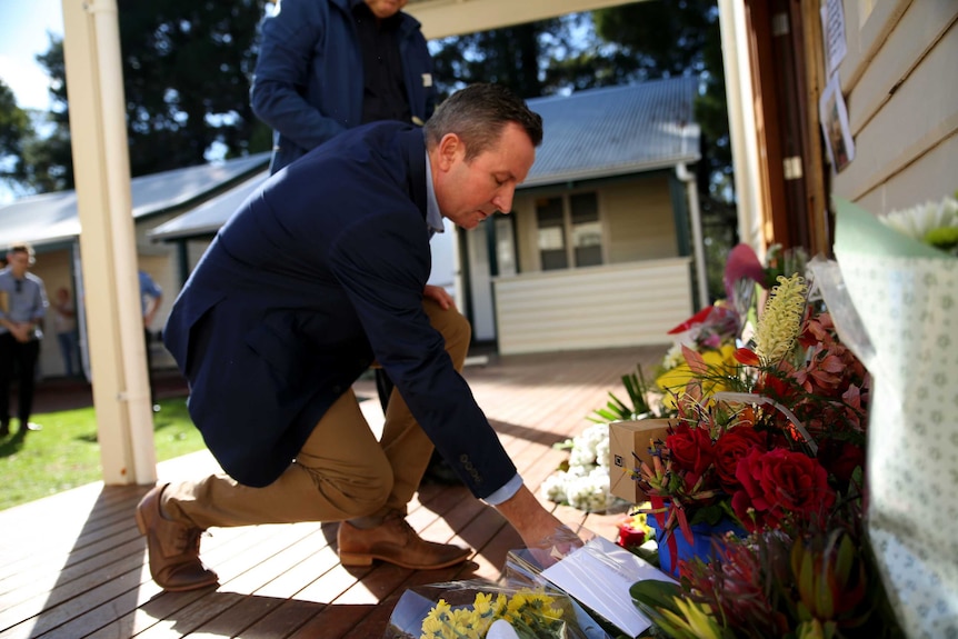 The premier crouches forward to place flowers along a wall where there are more flowers on a verandah.