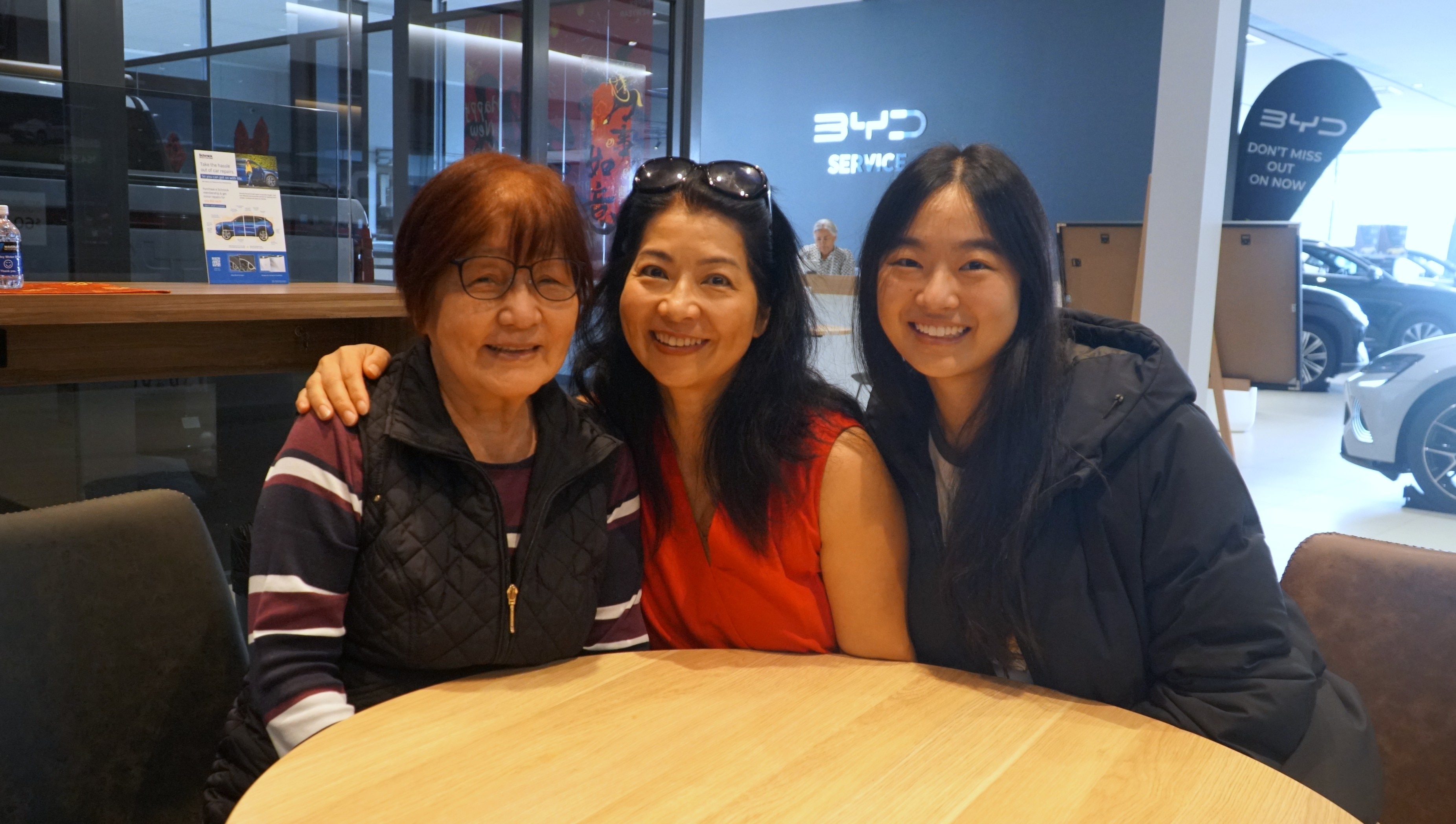 照片中，同一家族的三代女性围坐在汽车经销店的一张桌子旁。
