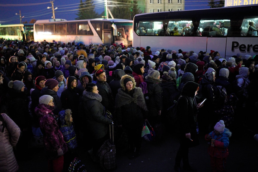 A large group of people in winter clothes stand huddled near a line of buses.
