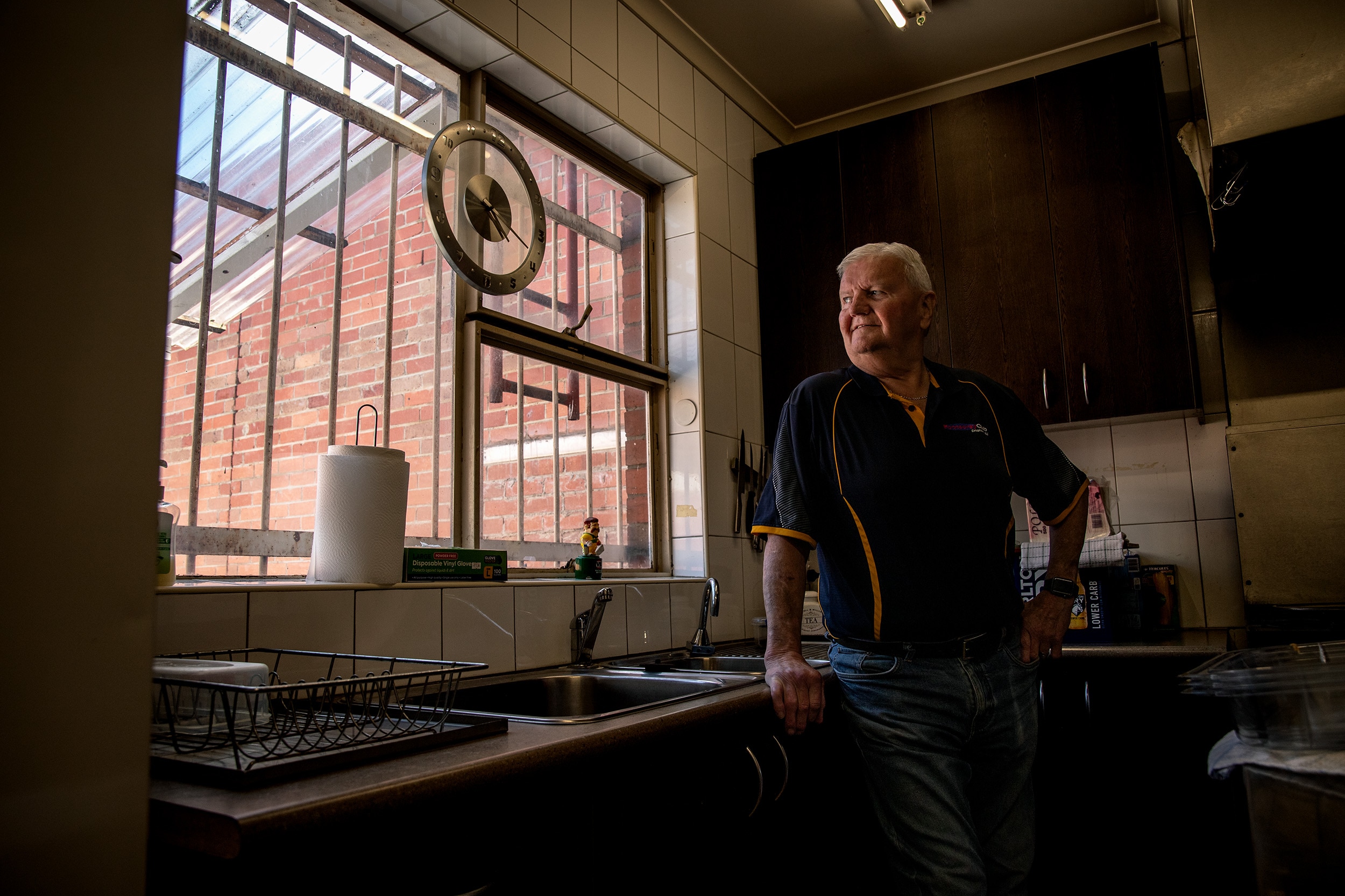 A man stands in front of a sink in a kitchen he is leaning on the bench and looking out a window behind him.