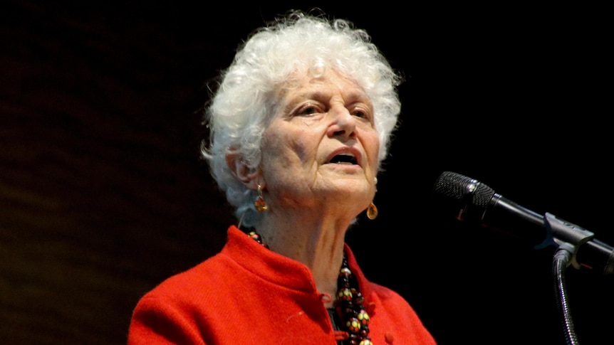 A mi-shot of Fiona Stanley wearing a red jacket speaking into a microphone against a black backdrop at an indoor forum.