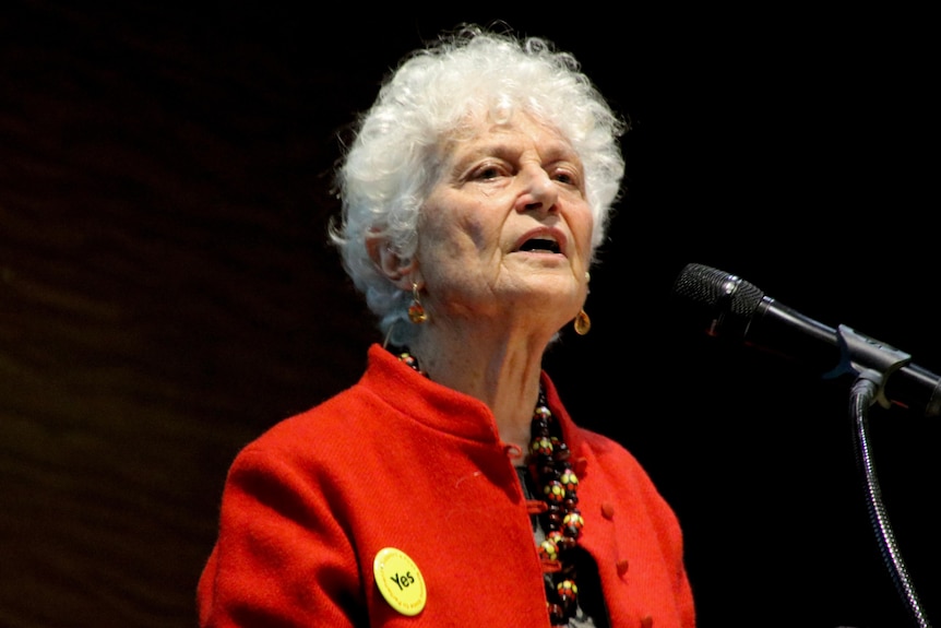 A mi-shot of Fiona Stanley wearing a red jacket speaking into a microphone against a black backdrop at an indoor forum.