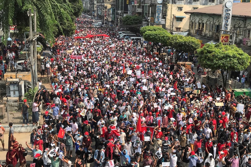 A huge crowd of people, many wearing red items of clothing,  line a main road waving red flags.