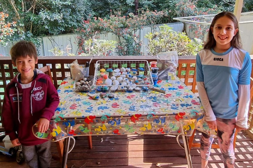 Two children stand in front of their painting table. 