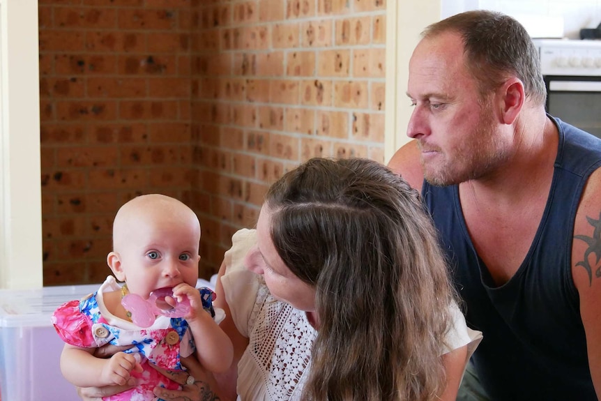 A woman and a man sit on the ground looking at a baby as she chews on a teething toy.