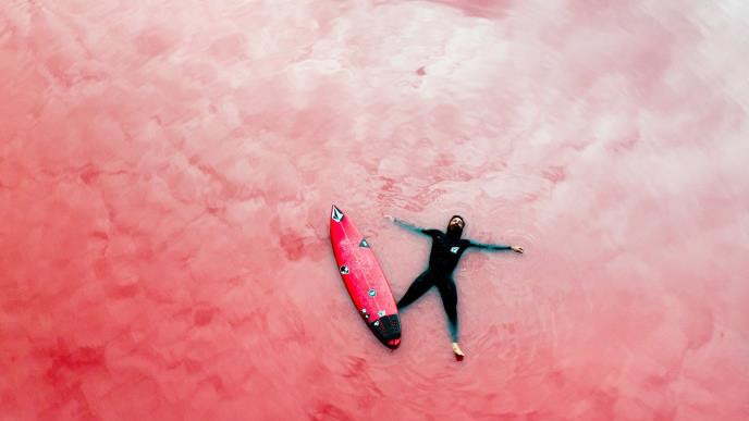 Bentley Dean, a bearded man in a wetsuit, floating next to his surfboard on a pink lake
