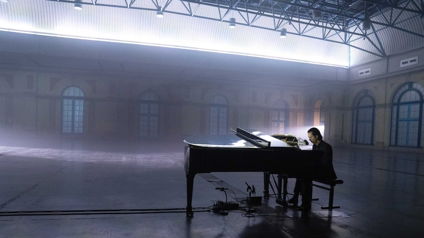 Nick cave playing a black grand piano in an empty warehouse