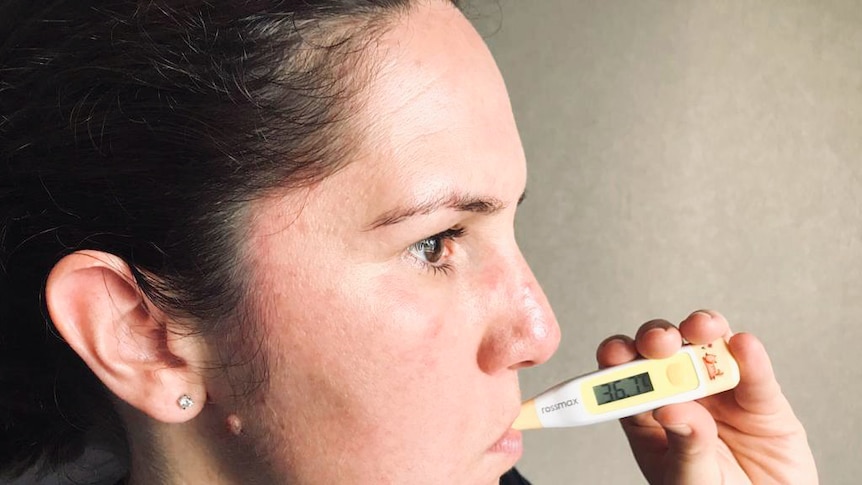 A woman with black hair holds a thermometer to her mouth as she faces off camera.